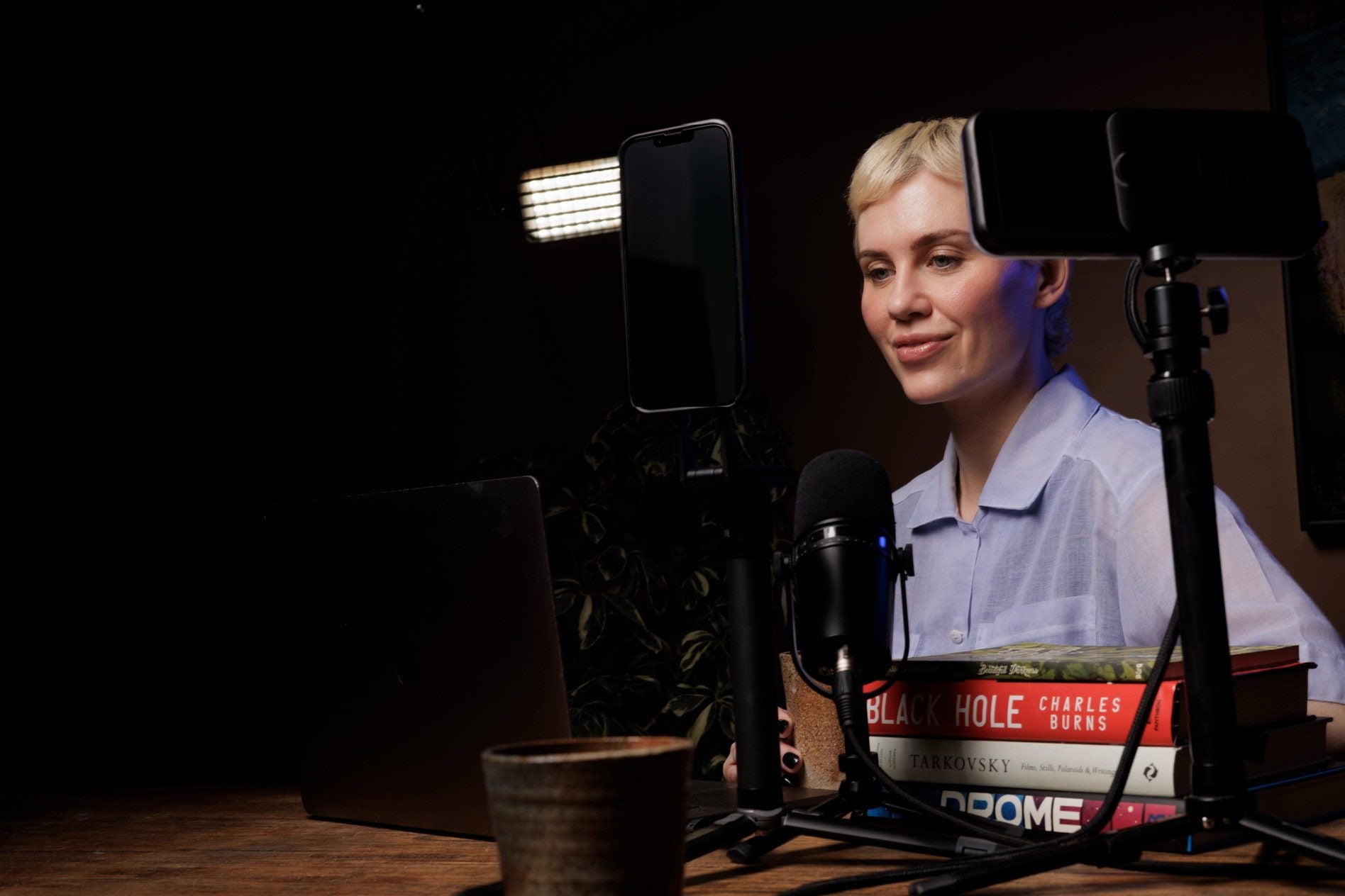 Lynda at a wooden desk, a microphone on a boom in front of her and a phone on a tripod at head height, warm light out of the black