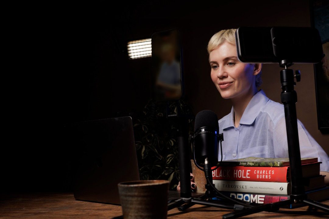 Lynda at a wooden desk, a microphone on a boom in front of her and a phone on a tripod at head height, warm light out of the black